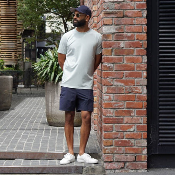 Model leaning against wall wearing Bound Pima Tee in Mint with Navy Shorts, Navy Cap, Sunglasses and White Sneakers